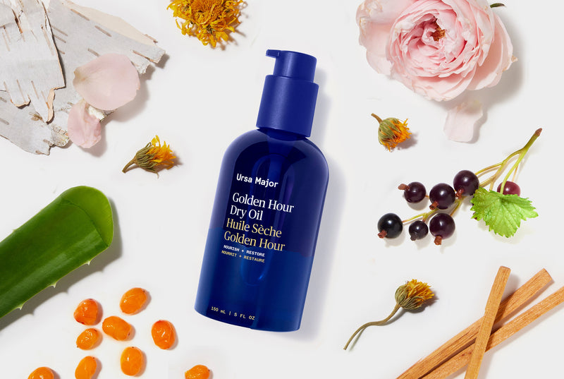 Blue bottle of Ursa Major Golden Hour Dry Oil surrounded by flowers and herbs on a white background